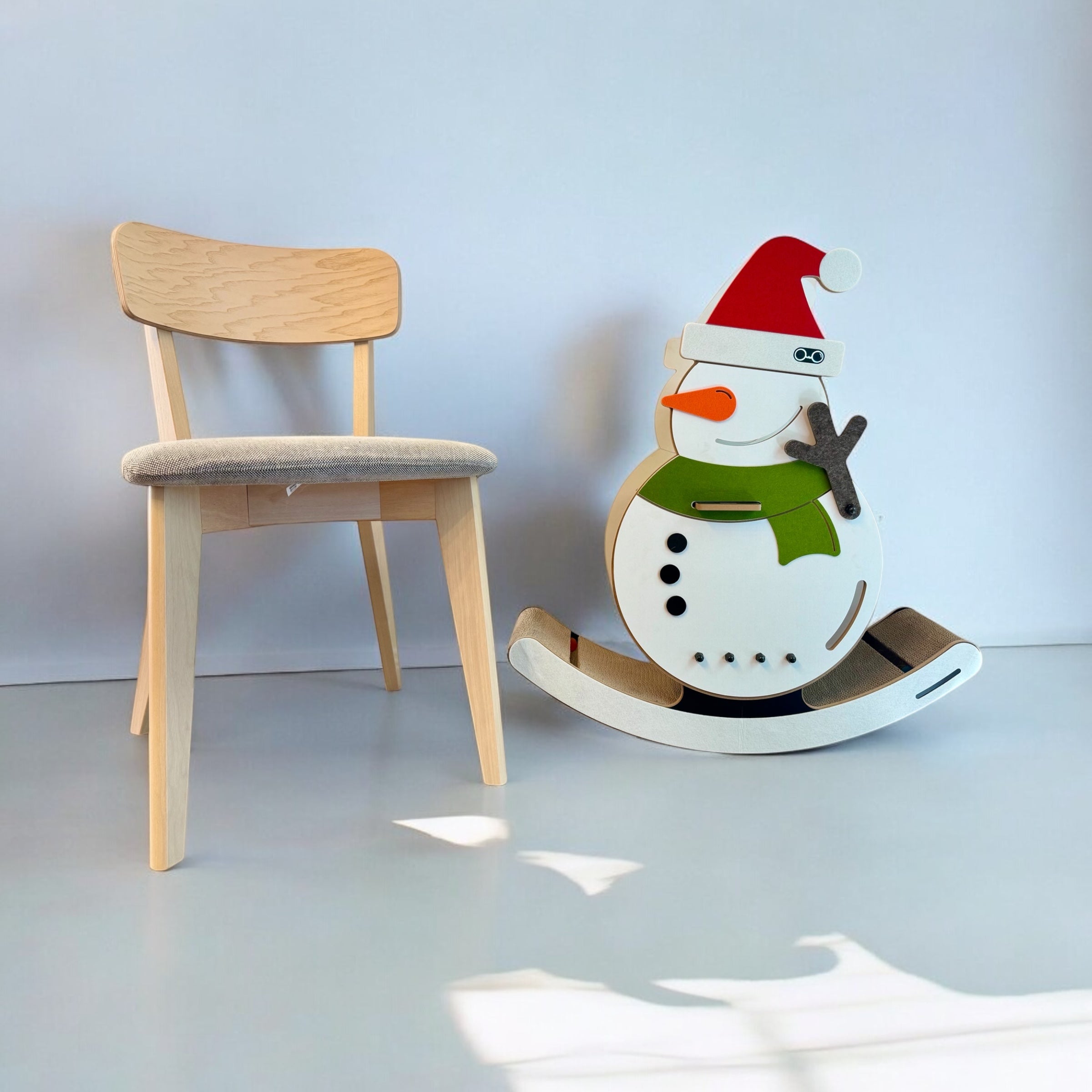 Lifestyle shot showing the scale of the large 6.6 kg snowman cat rocker placed next to a standard wooden dining room chair.